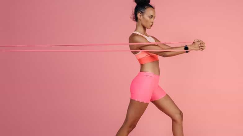 Woman doing a resistance band chest press against a pink background