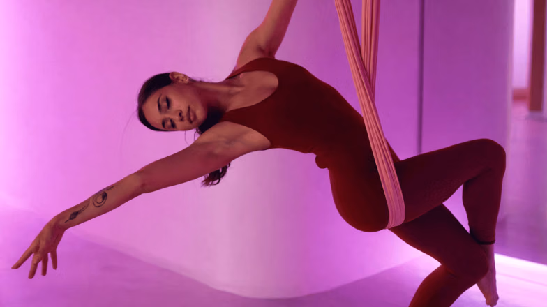 Woman performing an aerial stretching exercise on straps in a pink-lit fitness studio